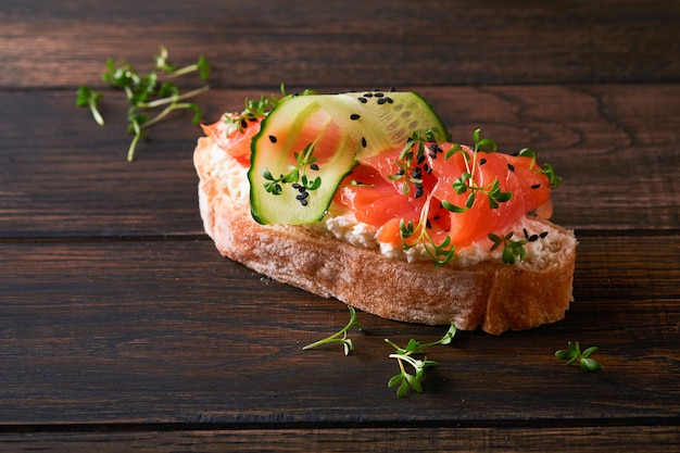 Sandwich salmone tostato con crema di formaggio cetriolo sesamo nero e microgreens sul vecchio tavolo in legno sfondo frutti di mare cibo sano fotografia in chiave bassa vista dall'alto