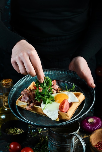 Sandwich with egg tomatoes and bacon on a plate in the hands of a chef On a black background Belgian waffles