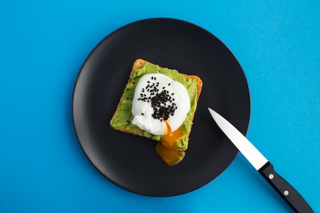 Sandwich with poached egg and avocado on the black plate in the center of the blue background