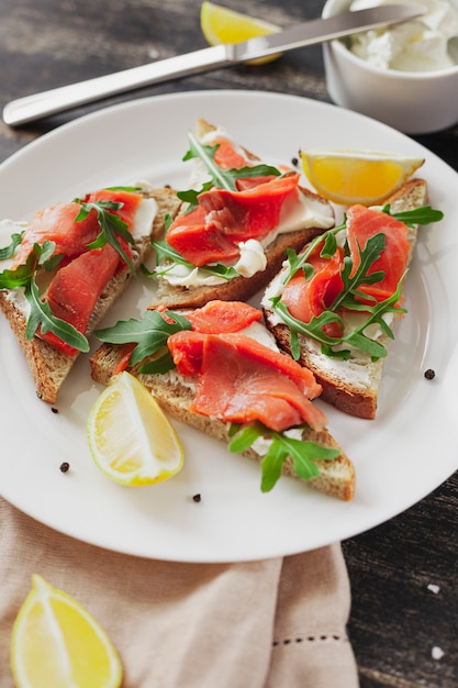 Sandwich with salmon, cheese, arugula and lemon on a white plate on a black table