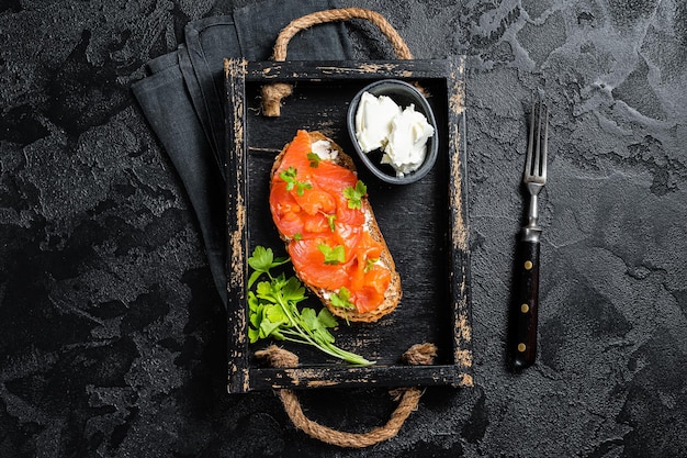 Фото Сэндвич с филе лосося и сливочным сыром в деревянном подносе черный фон вид сверху