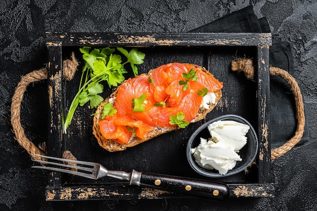 Sandwich with salmon fillet and cream cheese in wooden tray Black background Top view