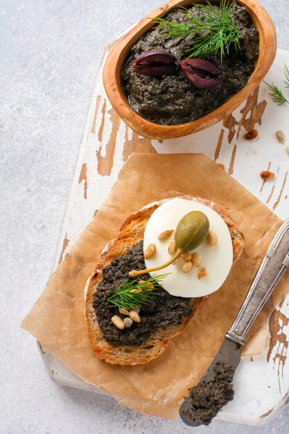 Sandwich with slice of mozzarella cheese and tapenade, caper on light grey rustic table background. Traditional Provence dish. Selective focus