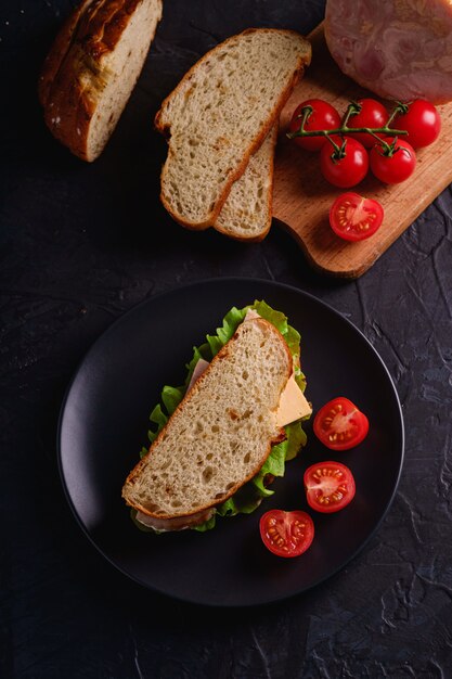 Sandwich with turkey ham meat, green salad, cheese and fresh cherry tomatoes slices on black plate near to ingredients on cutting board, dark textured wall, top view