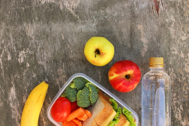 Sandwiches, fruits and vegetables in food box, water on old wooden background. Top view. Flat lay.