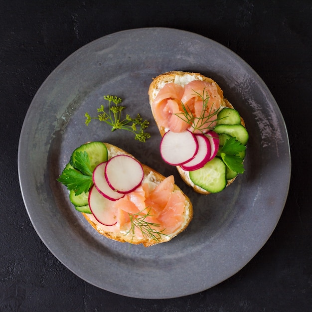 Sandwiches met gerookte roze zalm, radijs, komkommer en roomkaas op grijze keramische plaat en textiel oppervlak. Bovenaanzicht. Vierkant beeld