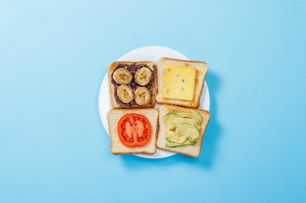 Sandwiches met kaas, tomaat, banaan en avocado op een witte plaat, blauwe ondergrond. Plat lag, bovenaanzicht.