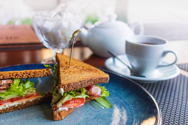 sandwiches met spek, kruiden en tomaten op een donkere plaat met een witte Kopje thee, een theepot en een suikerpot