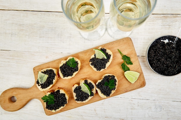 Sandwiches with black caviar. Sturgeon black caviar in wooden bowl, canapes and champagne on white background. Top view.