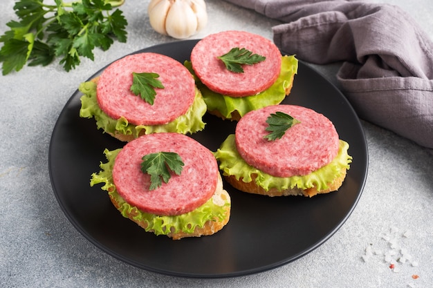 Sandwiches with lettuce leaves and sliced salami sausage on black plate.