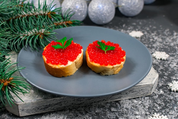クリスマスツリーとクリスマスボールの背景に石のテーブルの上に灰色の皿に赤いキャビアのサンドイッチ クリスマス・ニューイヤーズ・ホリデー 祭りの食べ物