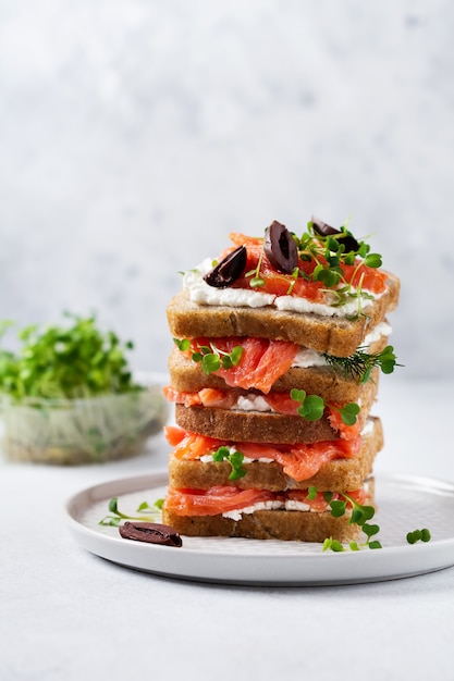 Бутерброды с копчеными оливками розового лосося, каламатой, микрогринами и сливочным сыром на серой керамической пластине и модном бетонном фоне. Традиционный скандинавский тост. Вид сверху.