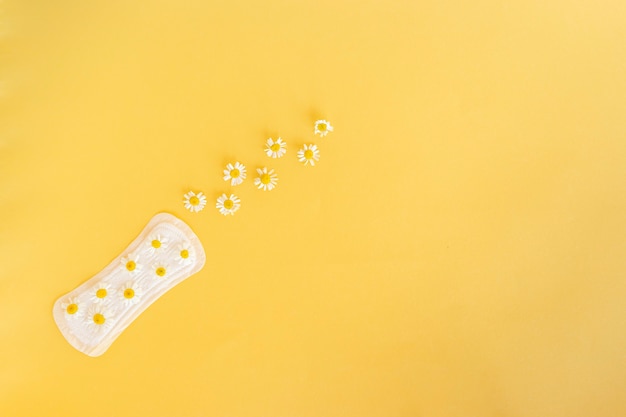 A sanitary pad with chamomile flowers on it lies on a yellow background. Menstruation days. Hygiene and freshness concept