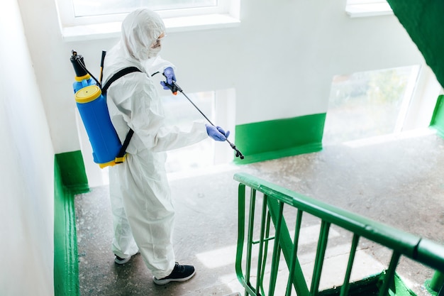 Sanitary professional worker wearing hazmat suit disinfects a staircase entryway block of flats. Coronavirus prevention measures at residential areas. 