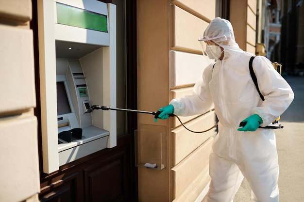 Санитарный работник опрыскивает банкомат дезинфицирующим средством во время пандемии коронавируса