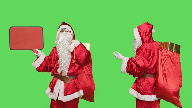 Santa character create advertisement using red speech bubble for marketing purposes, seasonal winter celebration. Father christmas showing empty billboard over greenscreen in studio.