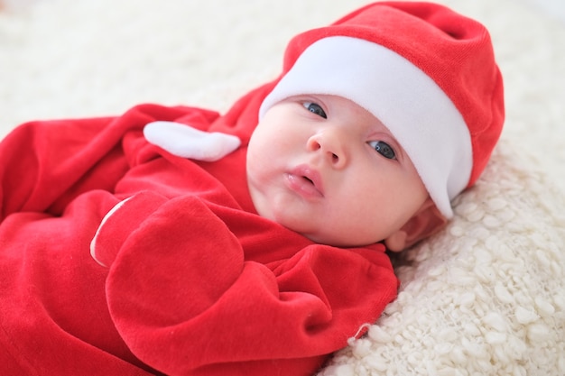 サンタの帽子で白い毛布のクリスマスの幼児の上に横たわっているサンタクロースの赤ちゃん。