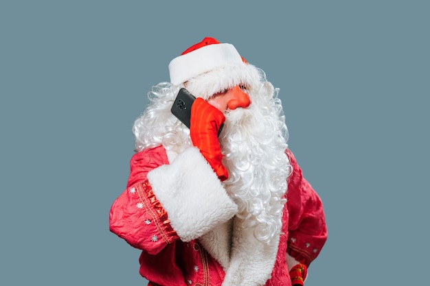 Santa Claus on blue studio background red hat white beard holds smartphone hand to ear speaks on it