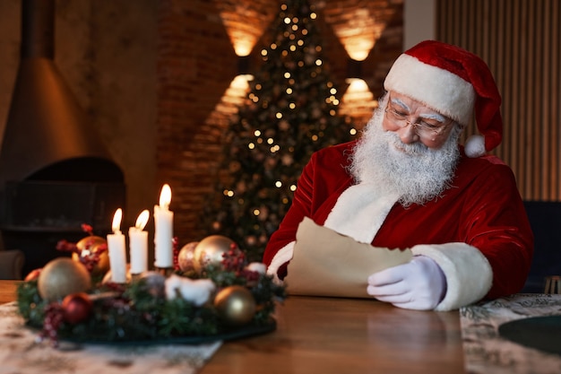 안경을 쓴 산타클로스는 크리스마스 cetrepiece와 함께 테이블에 앉아 어둡고 아늑한 공간에서 편지를 읽고 있습니다.