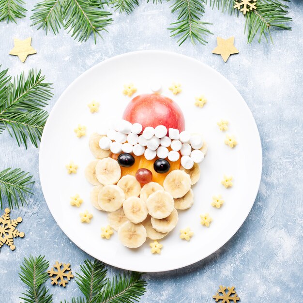 Лицо Санта-Клауса из фруктов и зефира на тарелке. Рождественские блюда для детей. Вид сверху