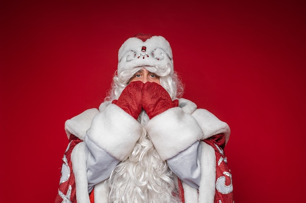 Санта-Клаус в причудливом традиционном костюме прикрывает рот обеими руками в красных рукавицах.