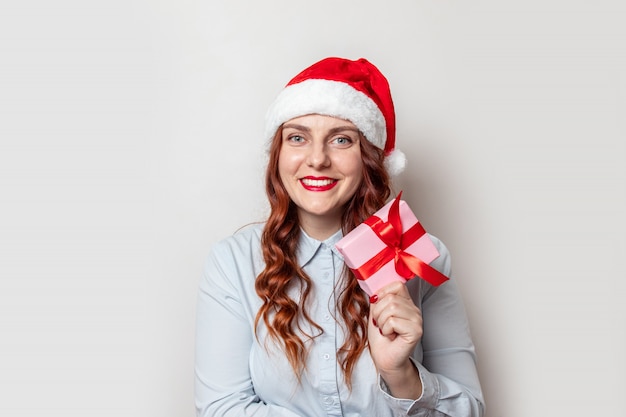 Santa Claus girl with curly hair and a red hat with a bumbon holds a gift box with a red satin ribbon bow and smiles on a  of gray wall. Merry Christmas and New Year web banner for site.