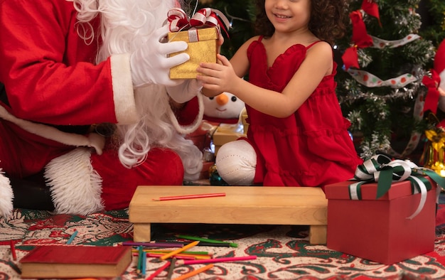 Santa Claus giving present to little cute girl near Christmas tree at home