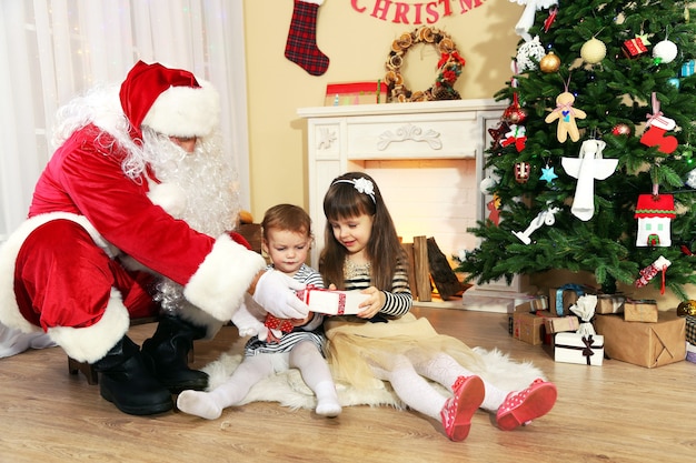 Babbo natale che fa un regalo a bambine carine vicino al camino e all'albero di natale a casa