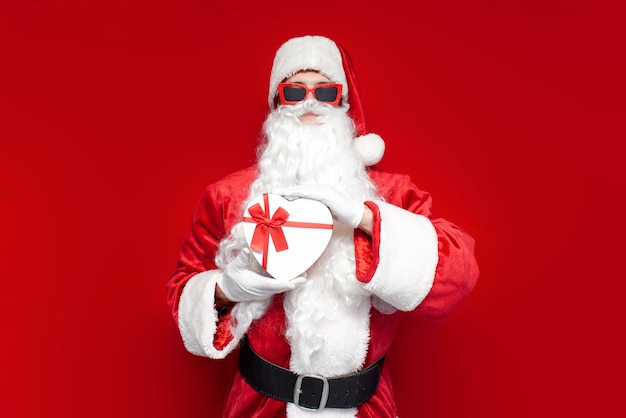 Santa claus in glasses holds gift box in the shape of heart on colored background man in hat