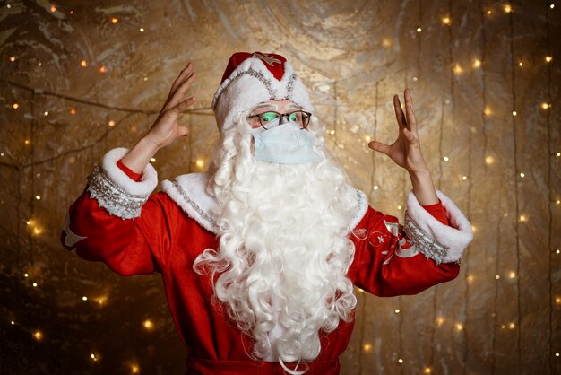 Santa claus in glasses and a mask against a virus shows a hand gesture against a wall background wit...