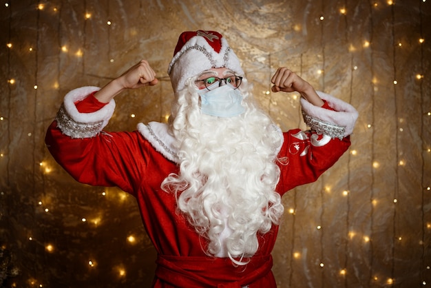 Santa claus in glasses and a mask shows gestures with his hands on the background of a wall with a g...