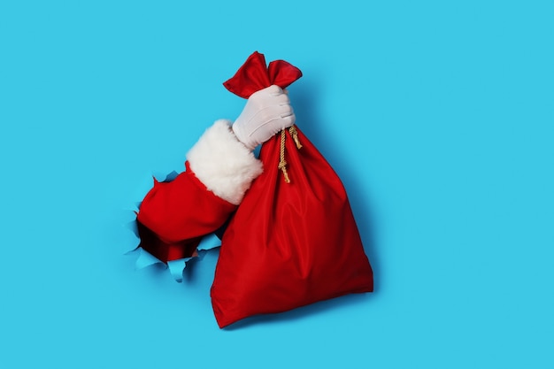 Photo santa claus hand holding red sack full of presents over light blue background