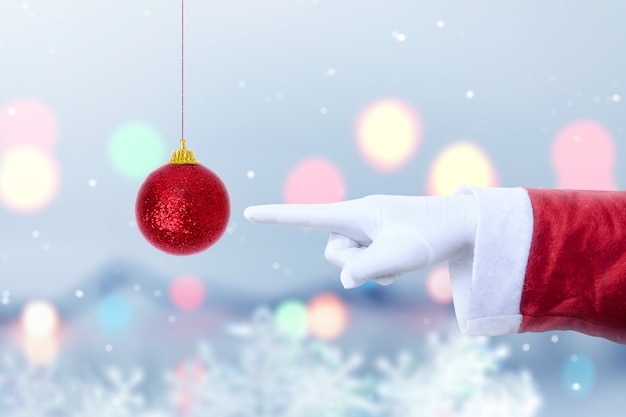 Santa Claus hand pointing red Christmas ball with blurred light background. Merry Christmas