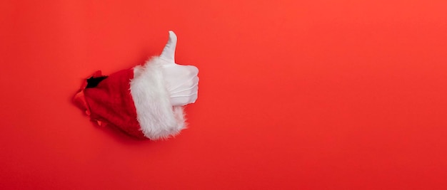 Рука Санта-Клауса поднимает палец вверх через дыру в красном бумажном фоне