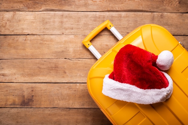 Santa claus hat lies on a yellow suitcase on a wooden background. Top view, flat lay.