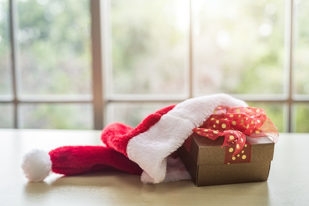 Шляпа Санта-Клауса с подарочными коробками, помещенными на деревянный стол интерьер комнаты, вид через окно с деревом Фон с копией пространства, украшения во время Рождества и нового года.