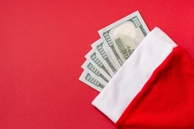 Шляпа Санта-Клауса с деньгами американских стодолларовых купюр на красном фоне, копией пространства