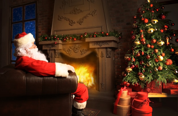 Santa Claus having a rest in a comfortable chair near the fireplace at home.