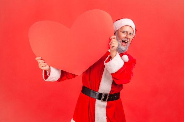 Санта-Клаус прячется за большим красным сердцем и смотрит в камеру с счастливым выражением лица.