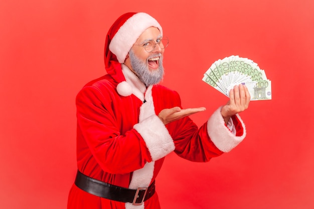 Santa claus holding fan of euro in hands, pointing at money, keeps mouth open.