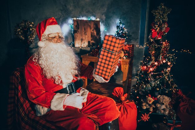 Санта-Клаус держит подарочную коробку дома Сядьте на стул, подумайте о плане отправки подарочной коробки для детей