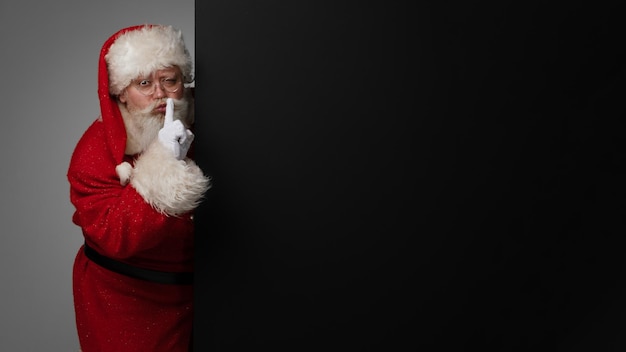 Santa Claus holding his finger on lips to have silence surprise concept, big black banner with copy space for text