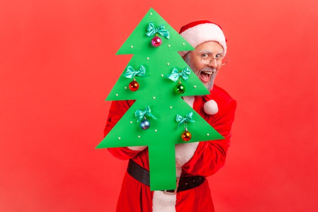 Santa claus holding paper christmas tree looking at camera with excited expression.