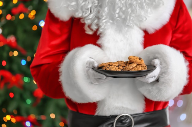 Santa Claus holding plate with cookies against blurred Christmas lights