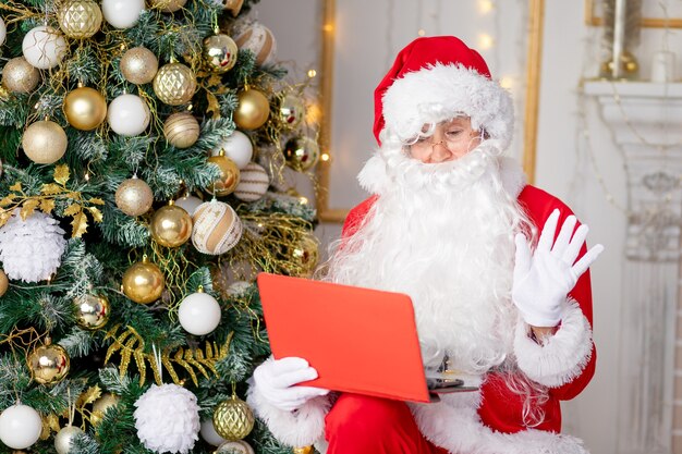 Santa Claus at home near the Christmas tree with a tablet c