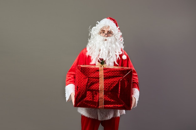 Santa Claus in glazen houdt in zijn hand het enorme kerstcadeau op de grijze achtergrond. .