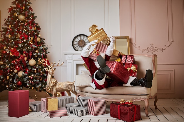 산타 클로스는 벽난로와 크리스마스 트리 근처에서 선물을 잔뜩 들고 소파에 누워 휴식을 취합니다. 새 해와 메리 크리스마스, 해피 홀리데이 개념