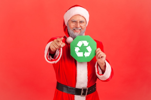 Санта-Клаус указывая на камеру пальцем, показывая зеленый знак утилизации.