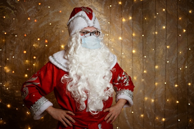 Santa claus posing in a mask on a wall background with a garland shows different gestures with his h...