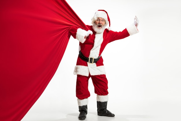 산타 클로스는 흰색 배경에 고립 된 크리스마스 선물로 가득 찬 거대한 가방을 당기고 있습니다. 전통 의상을 입은 백인 남성 모델. 2020년 새해, 선물, 휴일, 겨울 분위기. 귀하의 광고에 대 한 Copyspace입니다.